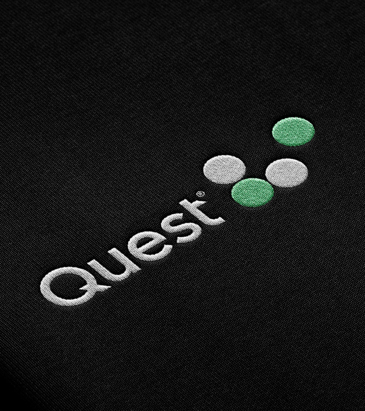 A close-up of the Quest logo embroidered on a black fabric surface, featuring three dots in white and green arranged in a triangular formation.