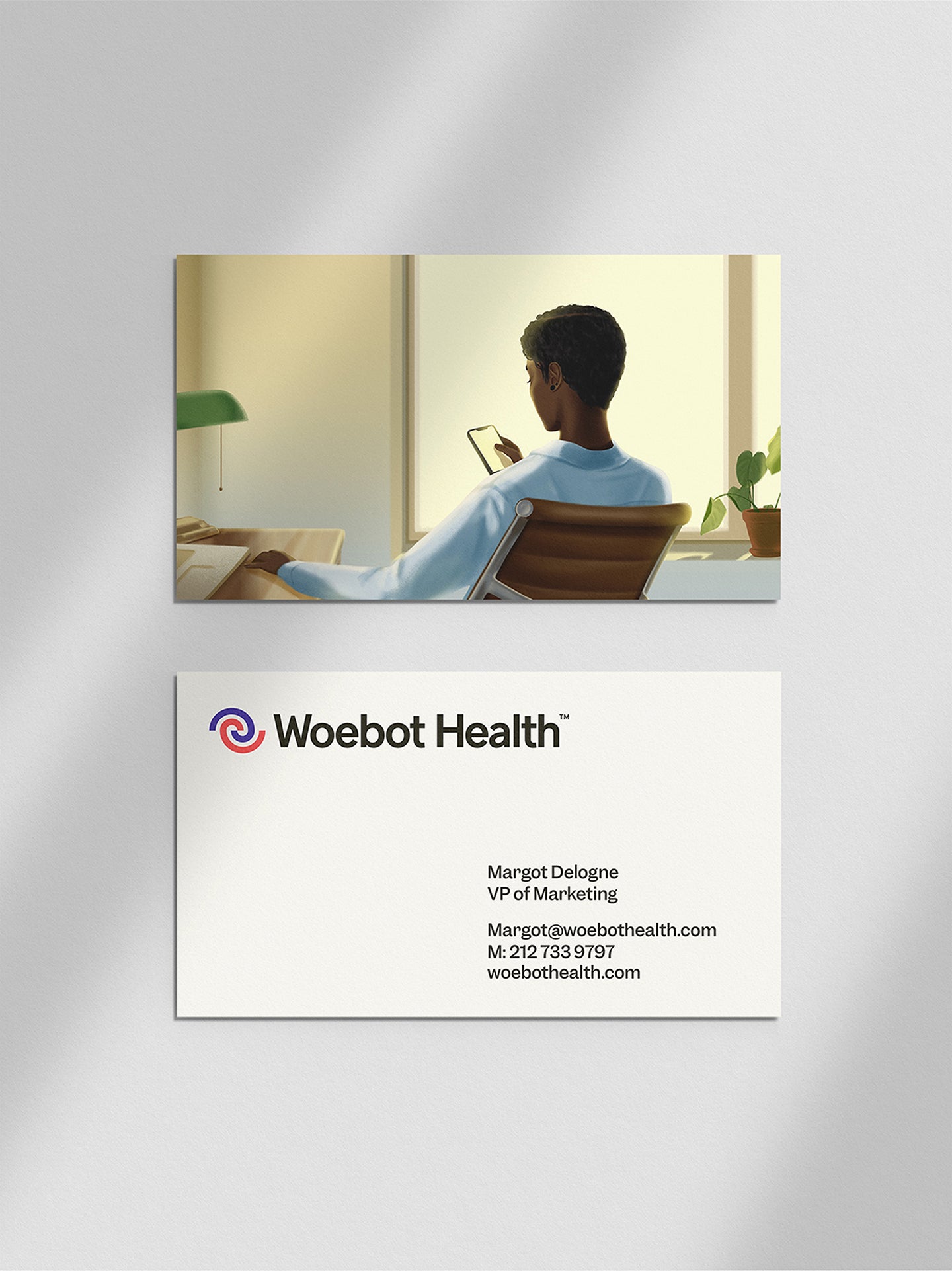 A Woebot Health business card displaying the name, title, and contact information of Margot Delogne, VP of Marketing. The back of the card features an illustration of a person sitting in a chair, looking at their phone in a well-lit room with a window and plants.