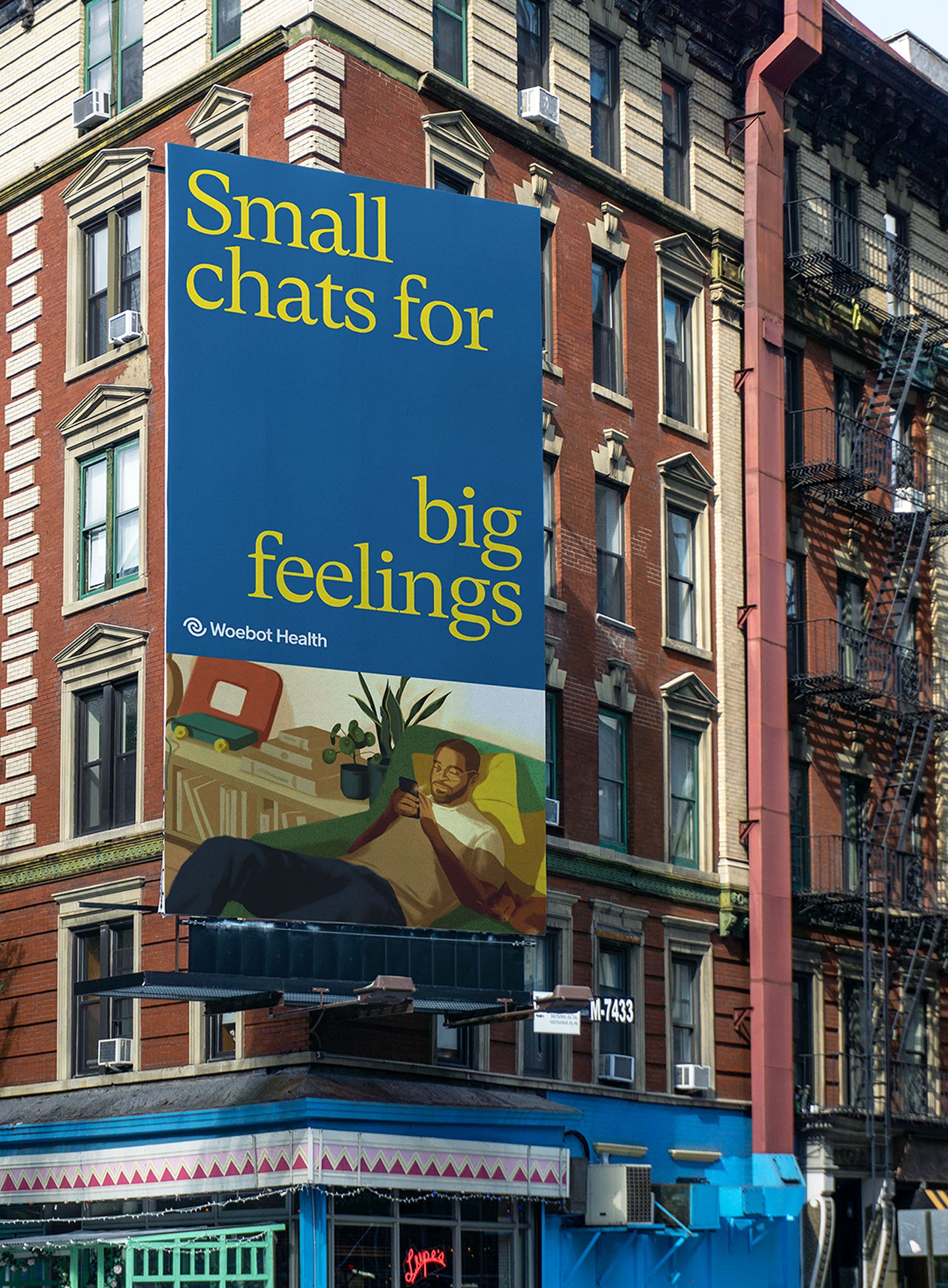 Large Woebot Health billboard on the side of a brick building, displaying the message 'Small chats for big feelings' in yellow text on a blue background. Below, an illustration shows a man lounging on a couch with his phone, highlighting the brand's mental health support services.