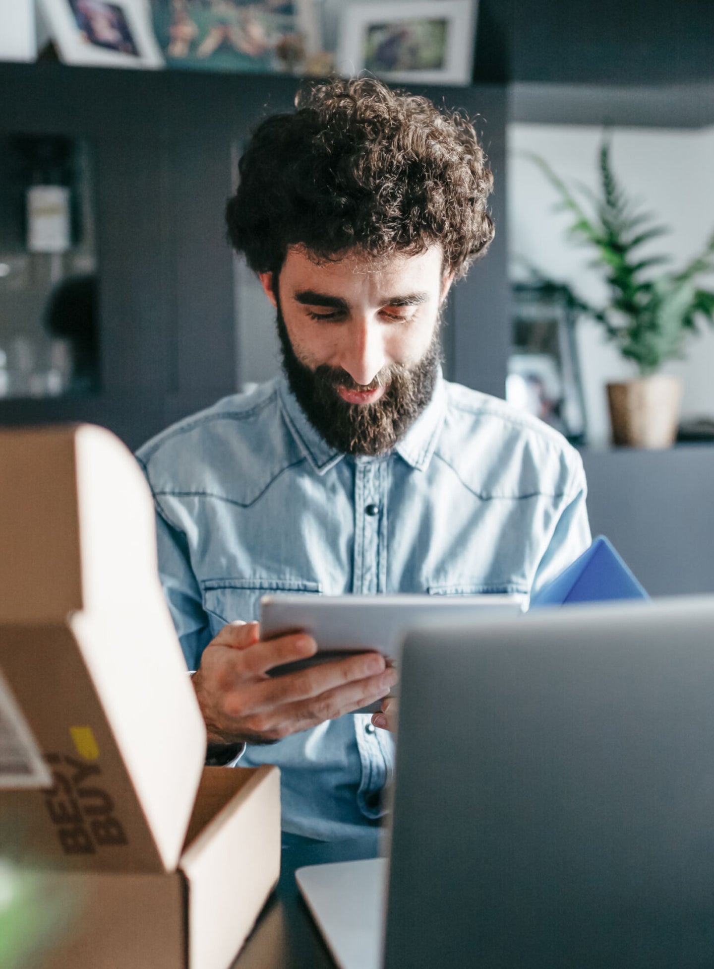 A bearded man in a denim shirt sits at a desk, looking at a tablet with a focused expression. An open Best Buy package and a laptop are in front of him, with a modern home setting in the background.