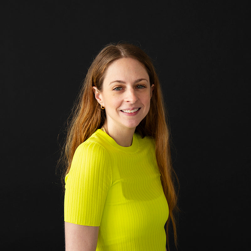 A portrait of a woman with long, light brown hair wearing a bright yellow short-sleeve top, smiling against a solid black background.