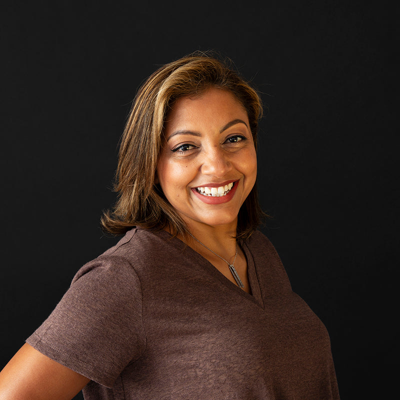 A portrait of a smiling woman with shoulder-length brown hair, wearing a brown V-neck top and a silver necklace, against a solid black background.