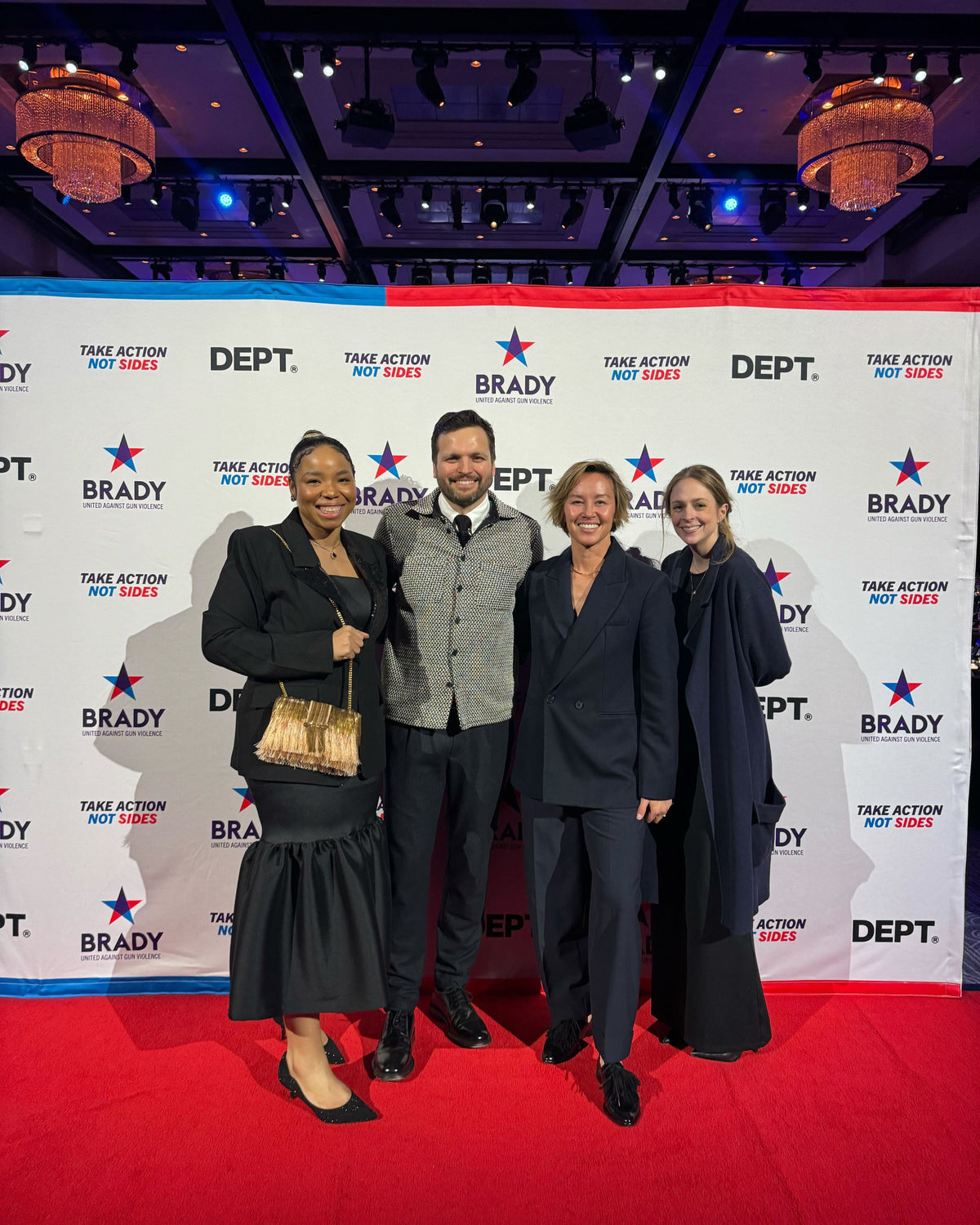 Four elegantly dressed individuals pose on a red carpet in front of a step-and-repeat banner featuring 'Brady,' 'Dept.,' and 'Take Action Not Sides' logos at a formal event.