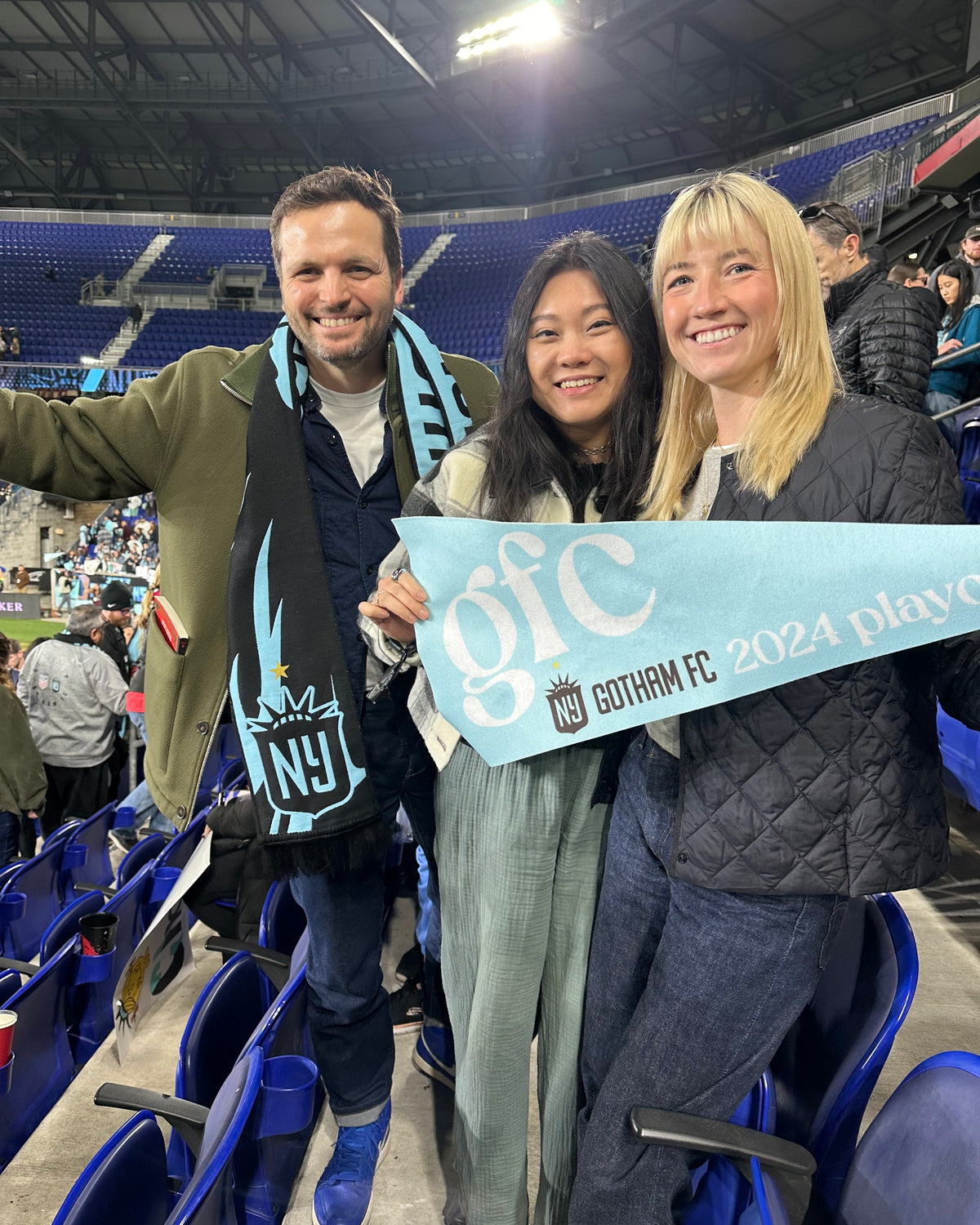 Three smiling fans stand in a stadium holding a light blue 'Gotham FC 2024 Playoffs' scarf. One wears a matching NY Gotham FC scarf, with a crowd and blue stadium seats in the background.