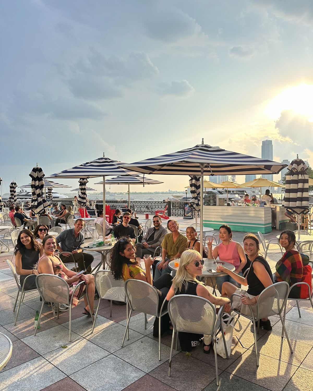 A large group of people gathers at an outdoor café with striped umbrellas, enjoying drinks and conversation during sunset. The setting overlooks a waterfront with a city skyline in the background.