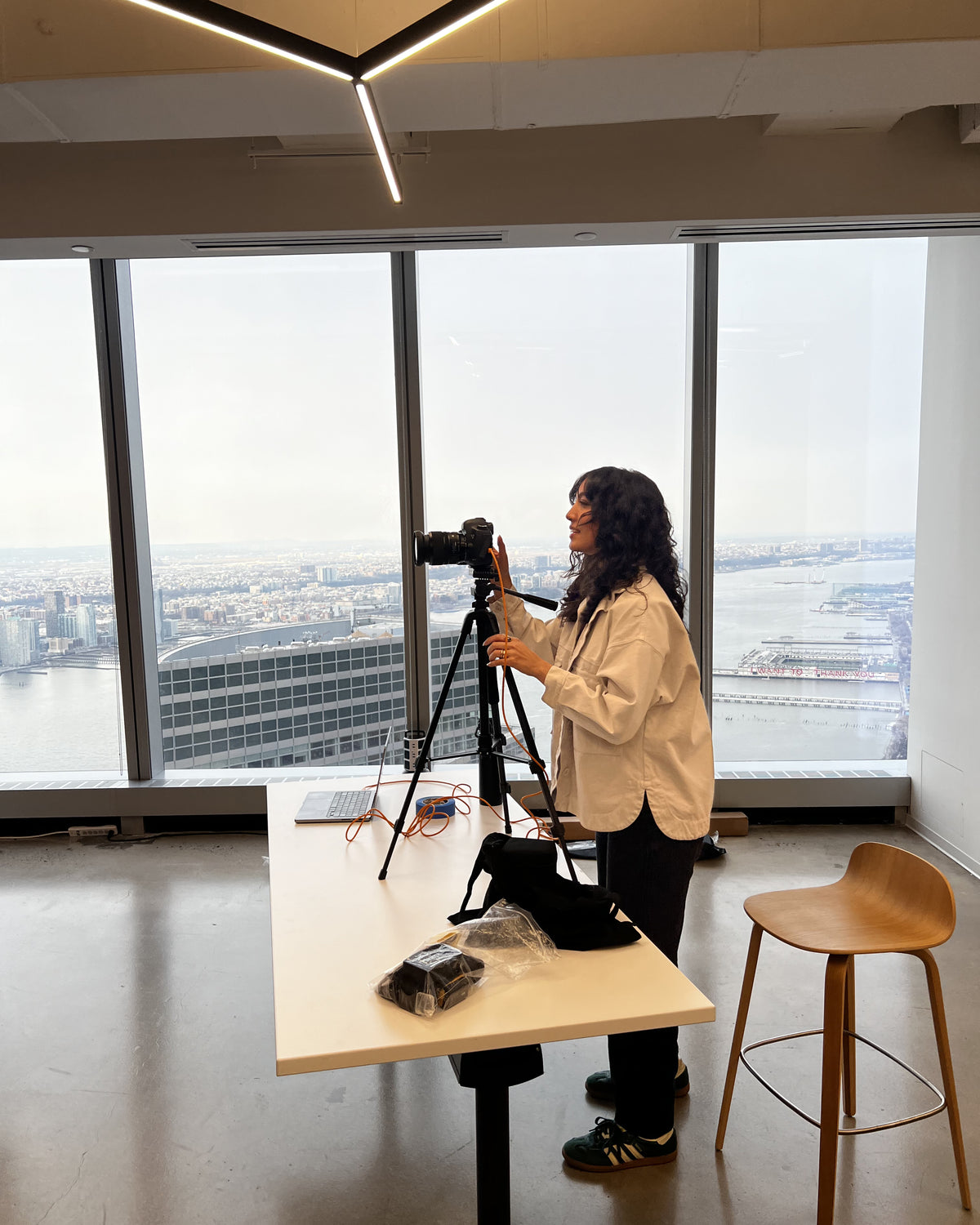 A woman in a white shirt sets up a camera on a tripod in a high-rise office with large windows overlooking a city skyline and waterfront. A table nearby holds a laptop and photography equipment.