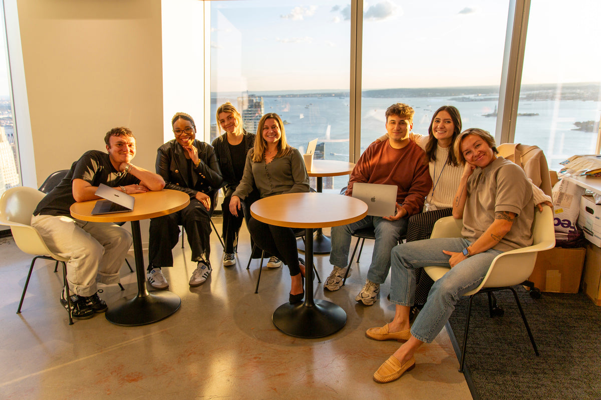 A group of seven colleagues sits around small tables in a modern office with floor-to-ceiling windows overlooking a waterfront. They are smiling and casually dressed, with laptops and notebooks nearby.