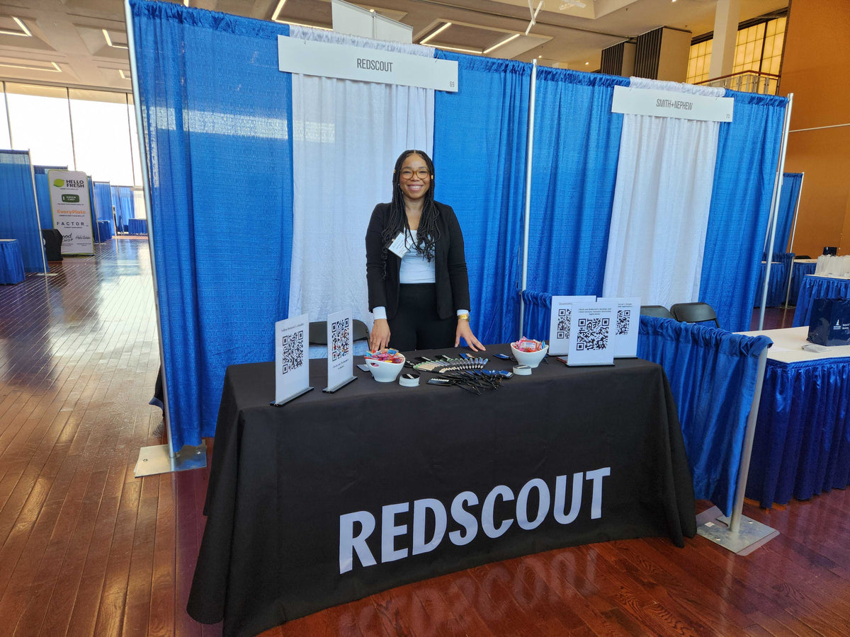 A smiling woman stands behind a REDSCOUT-branded career fair booth with promotional materials, QR codes, and small giveaway items on the table. Blue and white curtains form the booth backdrop in a bright event space.