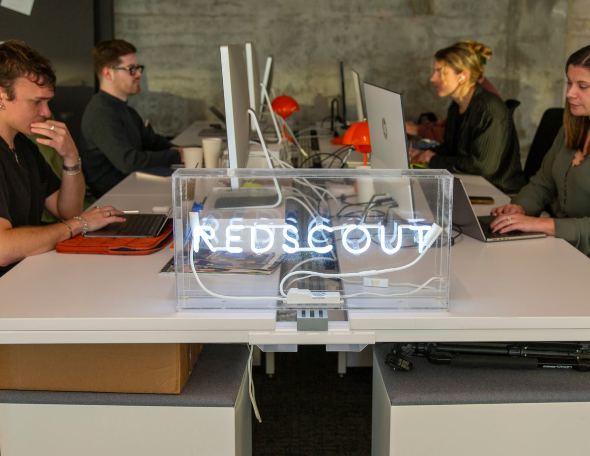 A modern office workspace with a group of four people working at a shared white desk. In the foreground, a neon sign encased in a clear acrylic box displays the word 'REDSCOUT.' Team members are focused on their laptops and desktop computers, with an industrial-style concrete wall in the background.