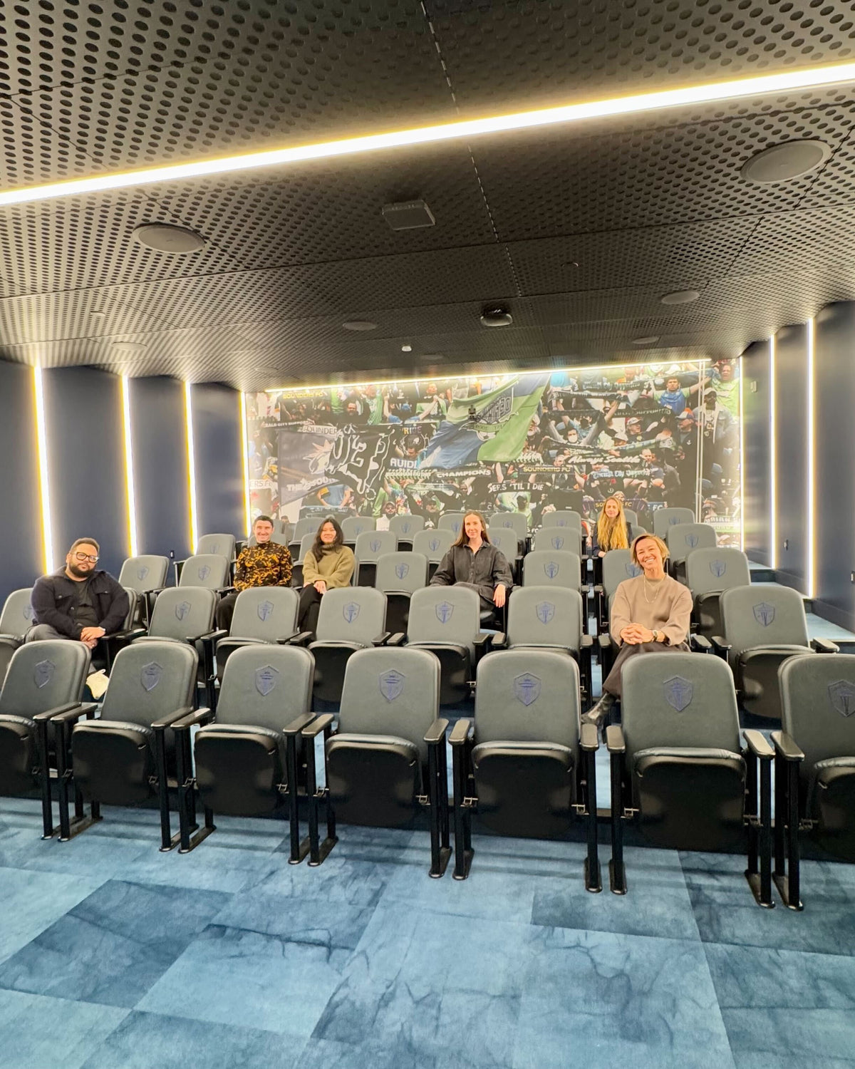 A group of six people sitting in a modern stadium-style meeting room with grey chairs embroidered with a shield logo. A large mural behind them displays a lively soccer crowd waving green and blue flags. The room features sleek lighting and a contemporary ceiling design.