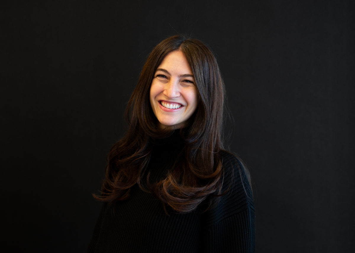 Portrait of a smiling woman with long, dark brown hair, wearing a black turtleneck sweater against a black background.