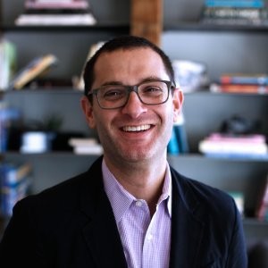 A man with short dark hair, glasses, and a smile, wearing a dark blazer over a light-colored shirt, standing in front of a bookshelf filled with books.