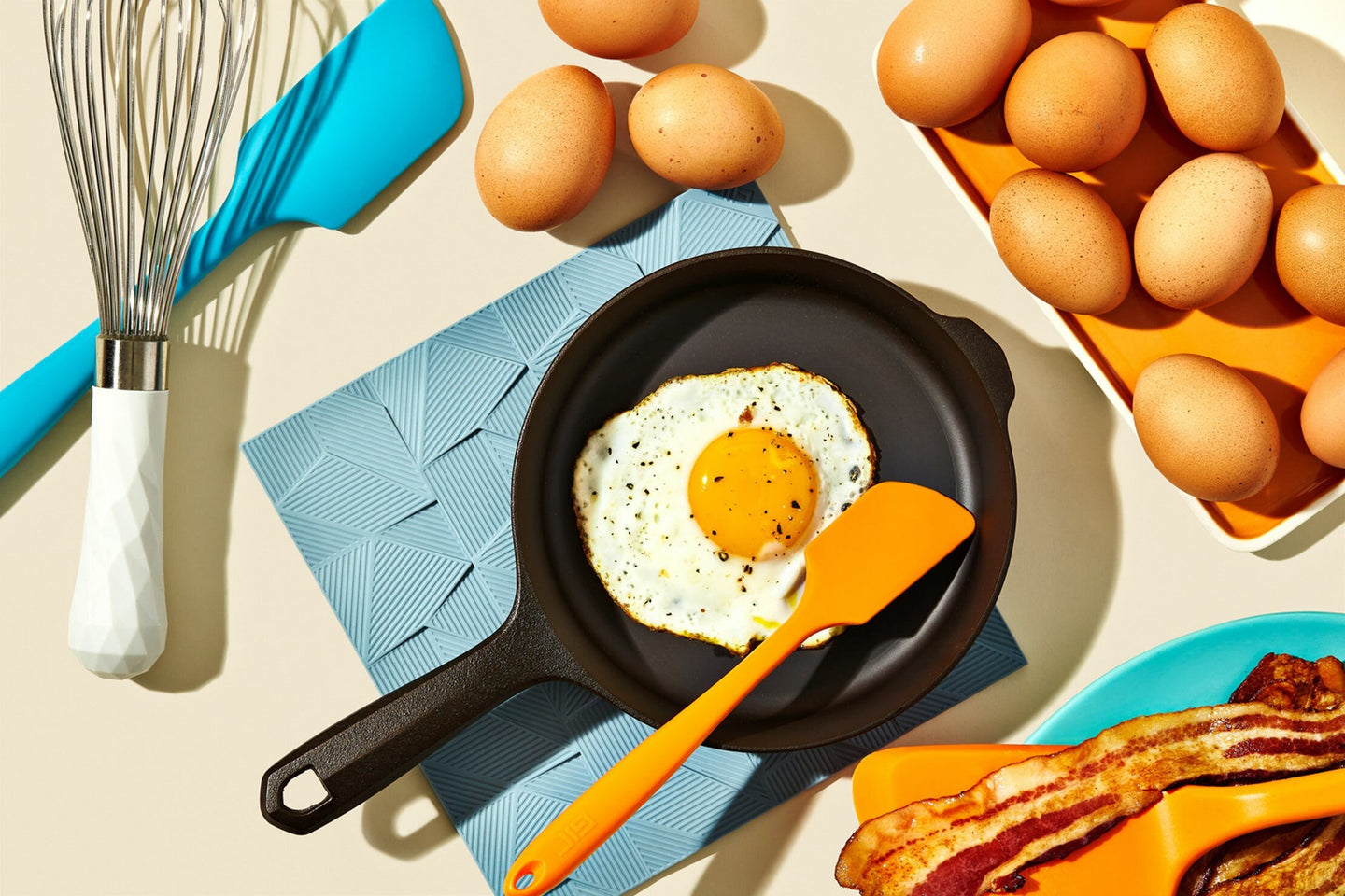 A top-down view of a frying pan with a sunny-side-up egg, surrounded by whole eggs, cooking utensils, and bacon.
