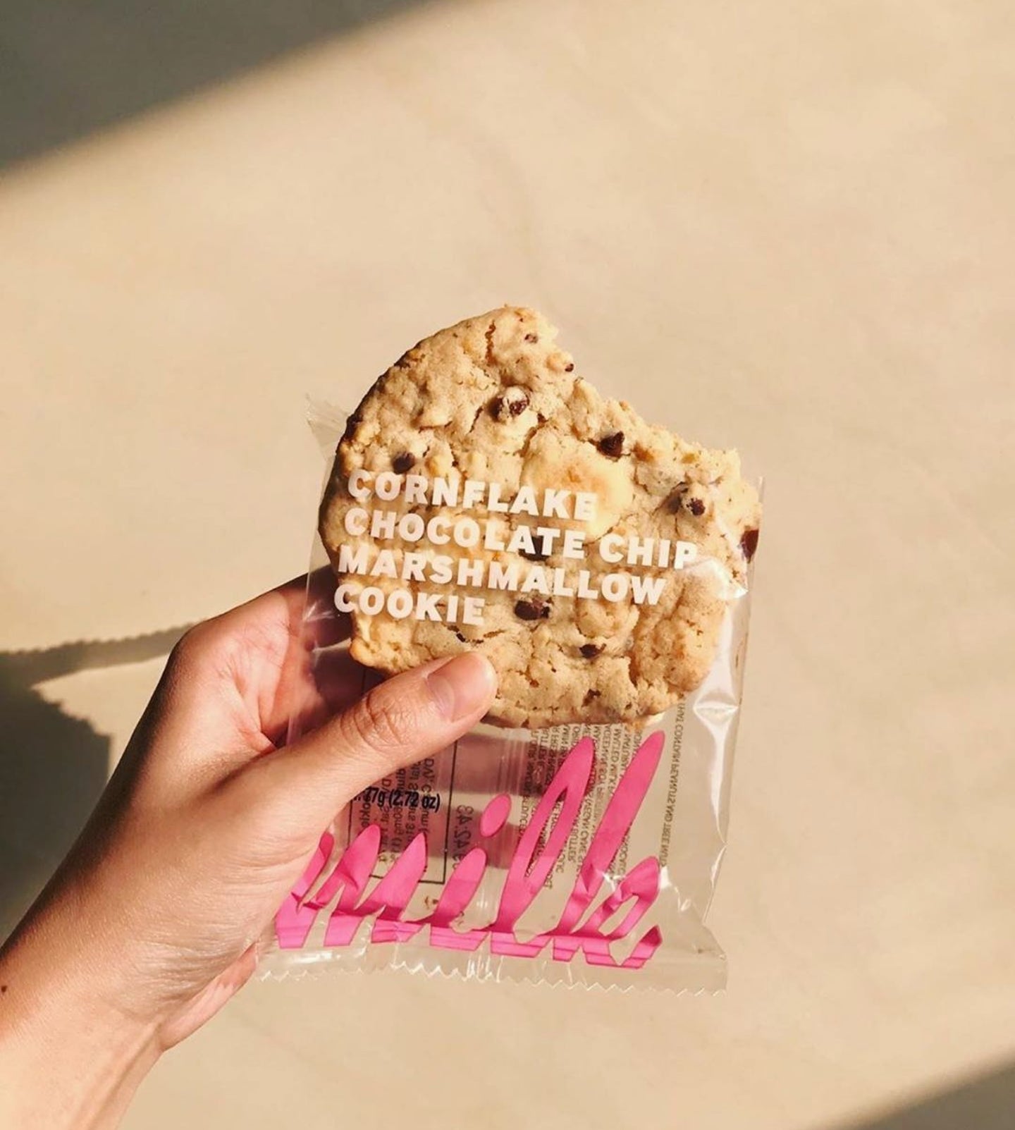 A hand holds a partially eaten Milk Bar Cornflake Chocolate Chip Marshmallow Cookie in its clear packaging with pink "milk" branding. The cookie is golden brown with visible chocolate chips and marshmallow bits. The background is softly lit with a warm beige tone.