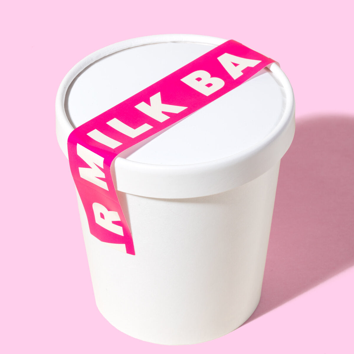 A white pint container sealed with a bright pink ribbon featuring bold white "MILK BAR" lettering, set against a pastel pink background.