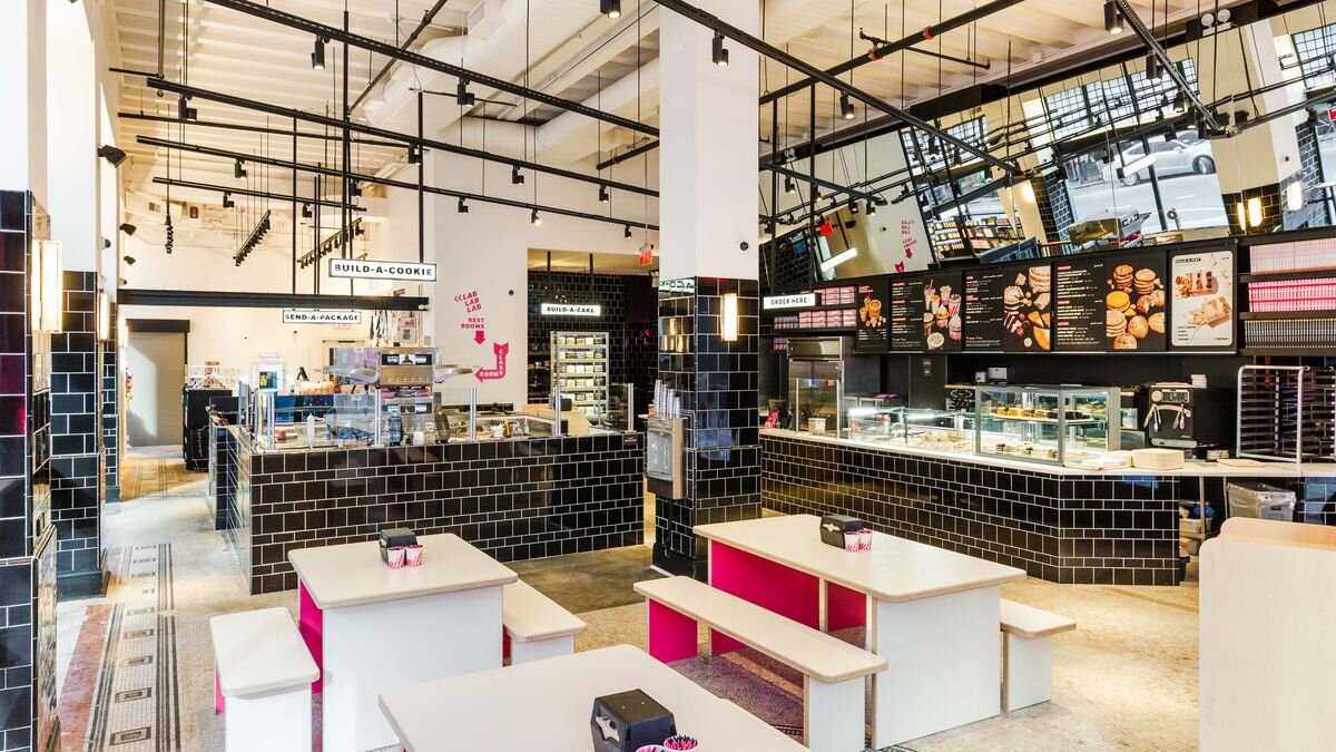 A well-lit, modern Milk Bar storefront with black tiled walls, white tables with pink benches, and digital menu boards showcasing baked goods and desserts.