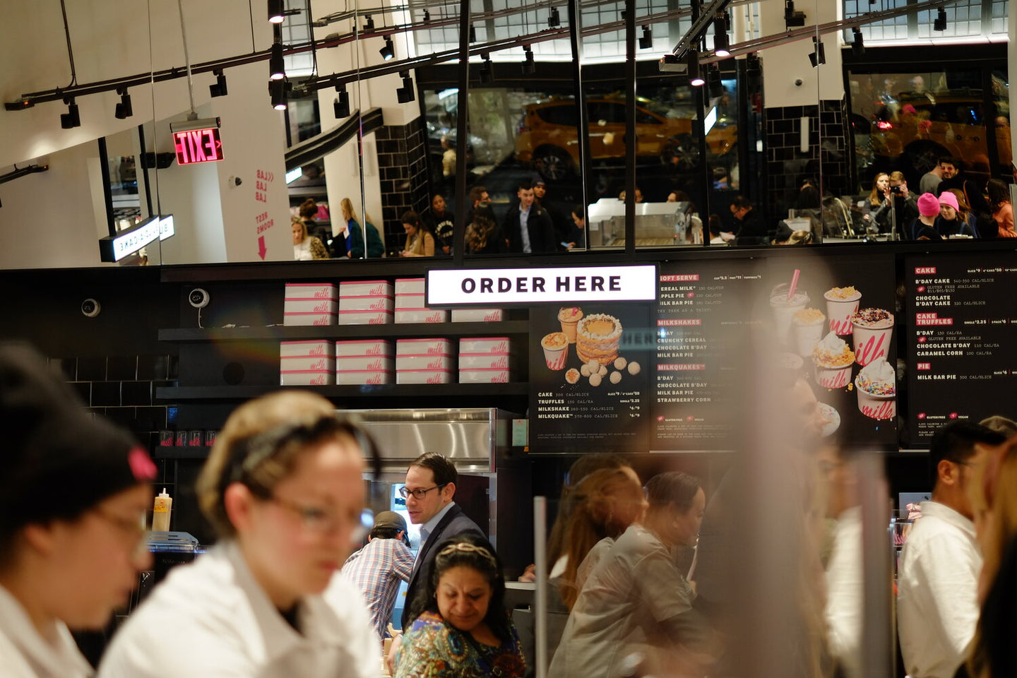 A bustling Milk Bar store interior with an "ORDER HERE" sign above the counter, reflective mirrors, stacked pink-branded boxes, and customers in the background.