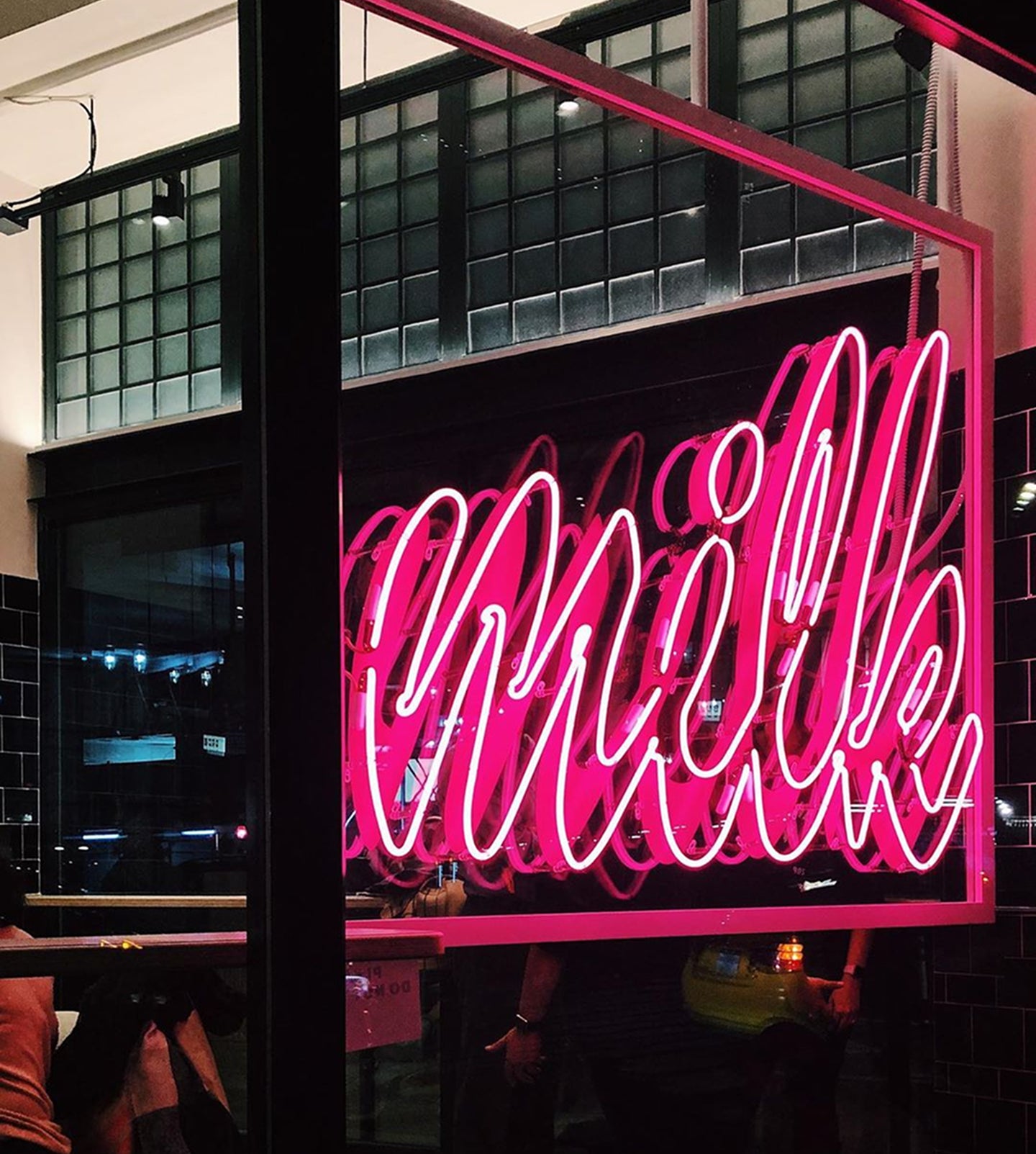 A glowing pink neon "Milk" sign inside the Milk Bar store, reflecting off large industrial-style windows.