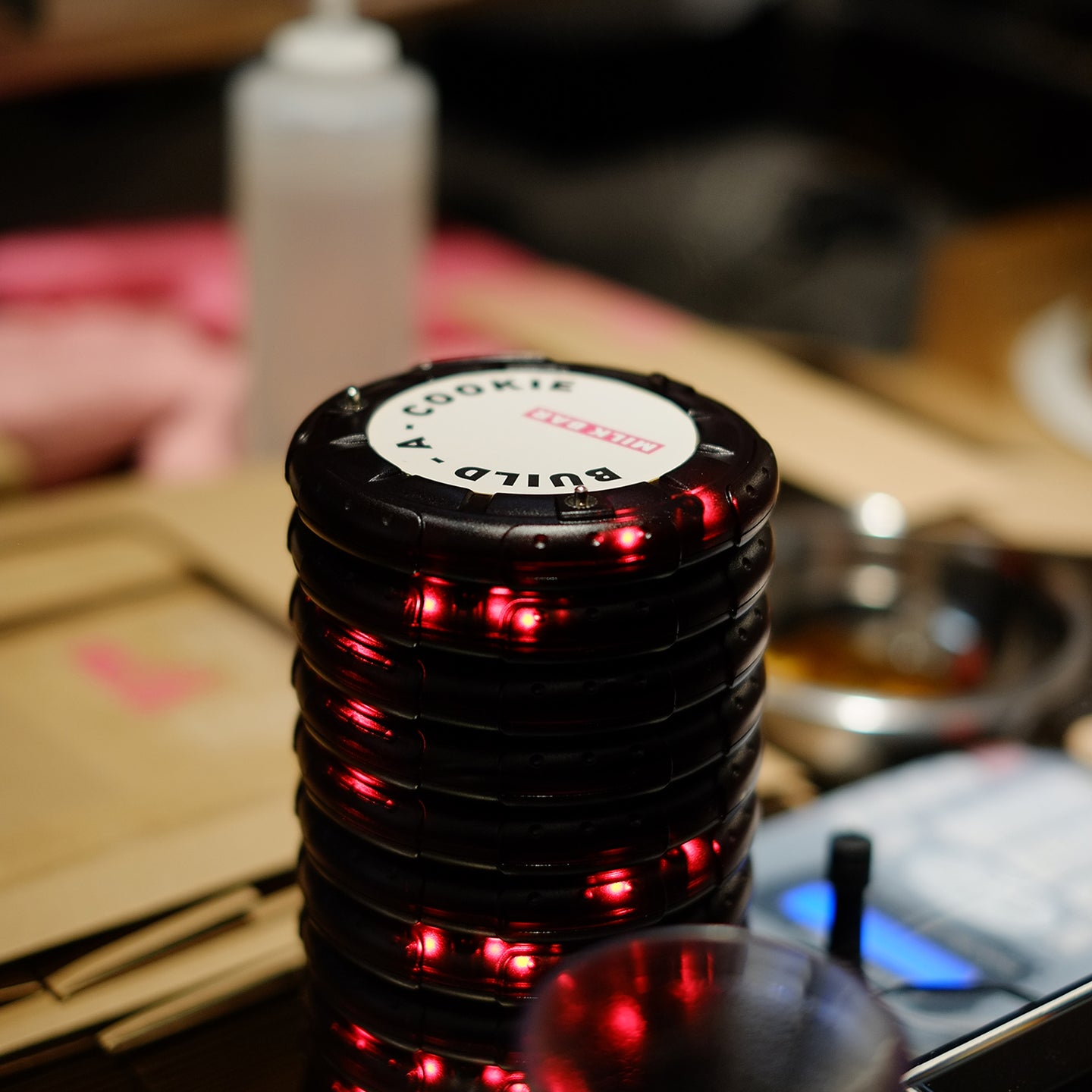 A stack of black buzzers with red LED lights, labeled "Build-a-Cookie," resting on a countertop with blurred kitchen elements in the background.