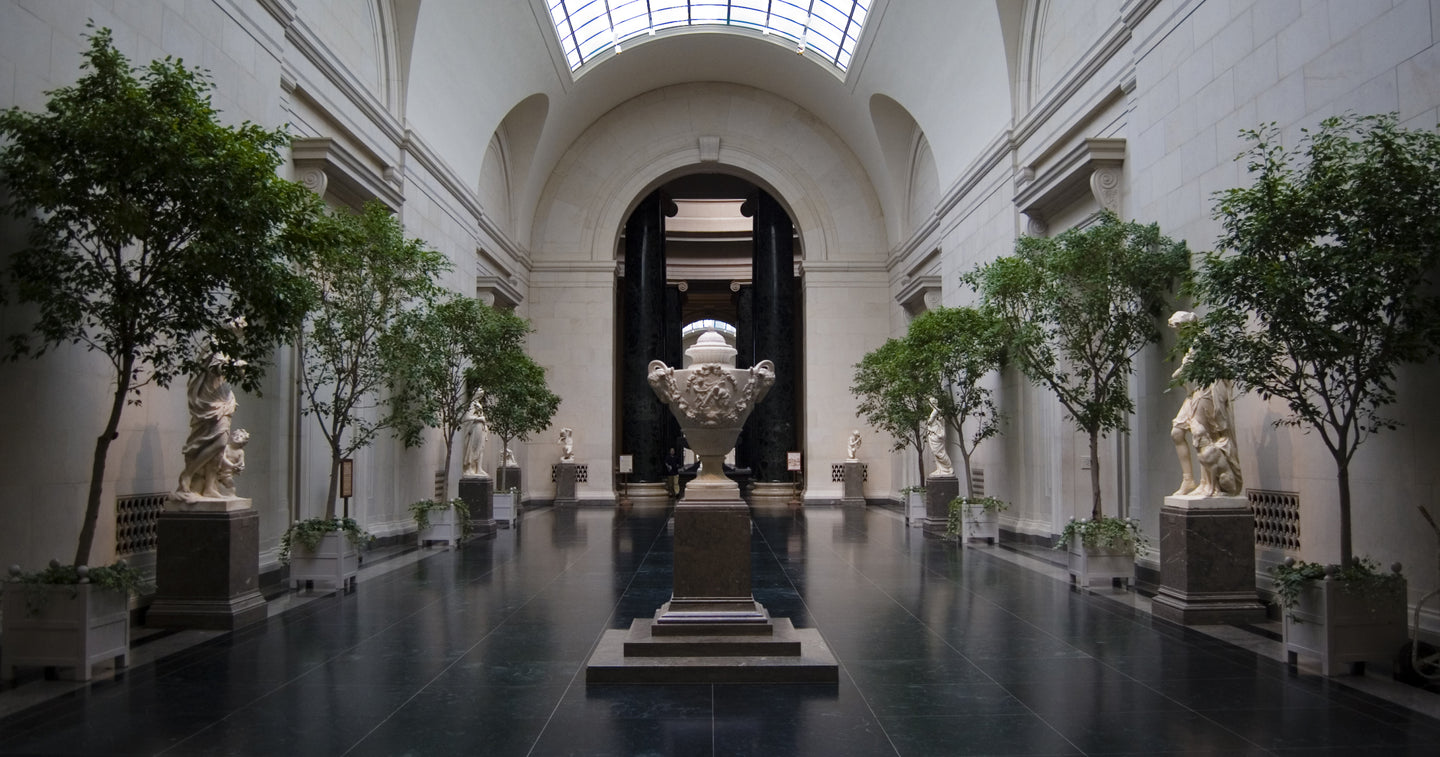 Grand museum hall with high arched ceilings, a skylight, and a symmetrical arrangement of trees, statues, and a central ornate urn on a pedestal.