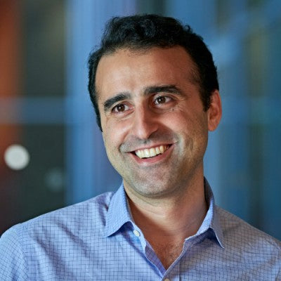 Portrait of a smiling man with short dark hair, wearing a light blue checkered dress shirt, standing in a softly lit indoor environment with a blurred background.