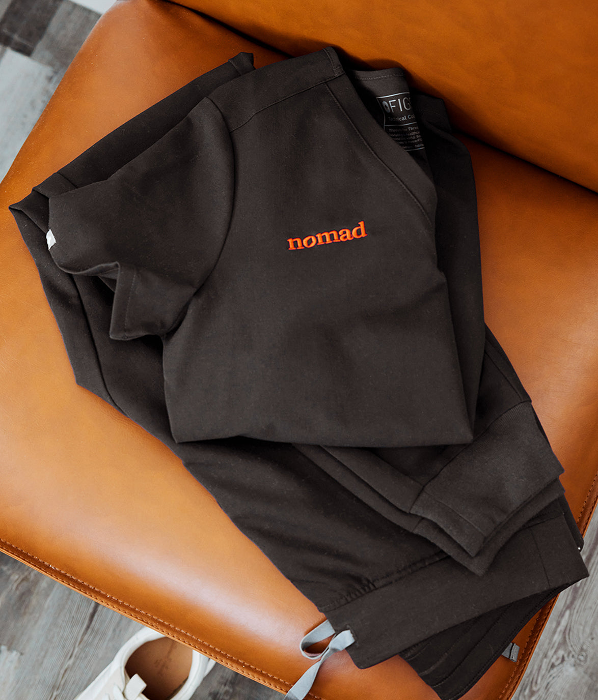 Folded black medical scrubs with the 'Nomad' logo embroidered in orange, placed on a brown leather chair.