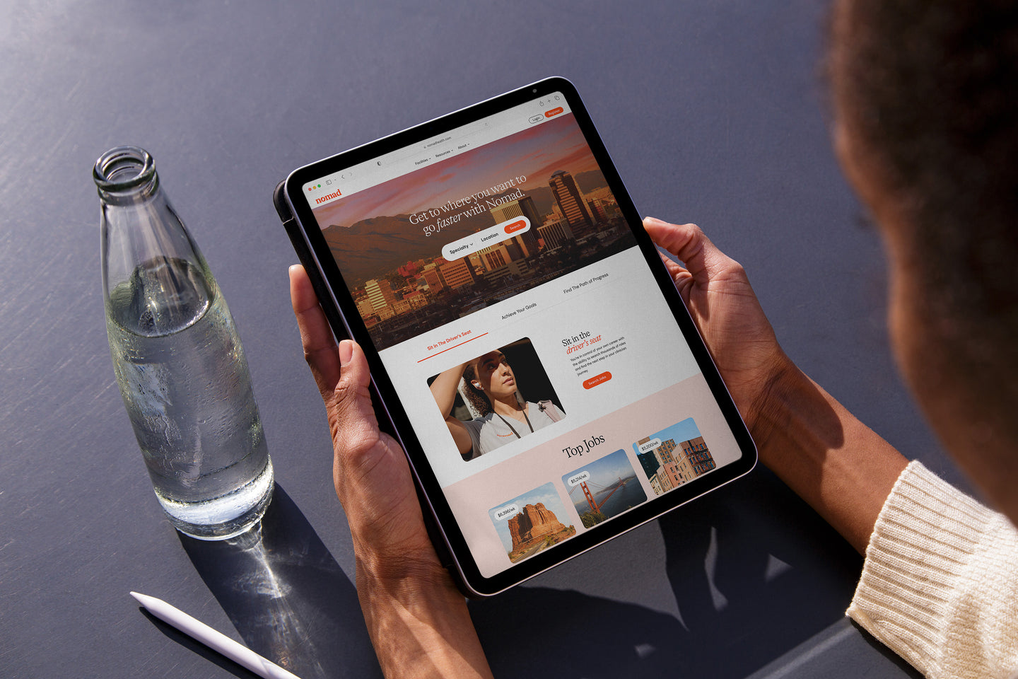 Person holding a tablet displaying the Nomad website, which features job opportunities and a banner with a scenic city view. A water bottle and stylus rest on the table nearby.