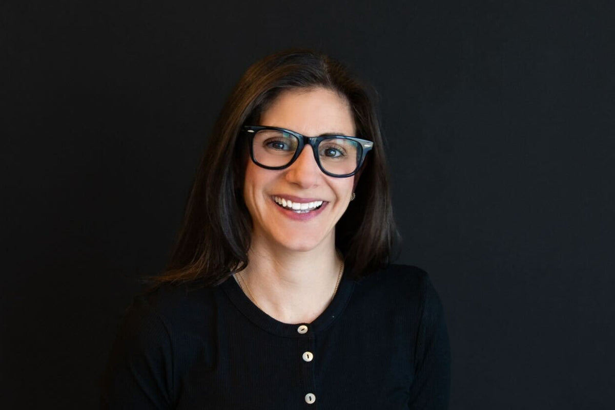 A smiling woman with dark hair and glasses wearing a black long-sleeve button-up top, standing against a solid black background.