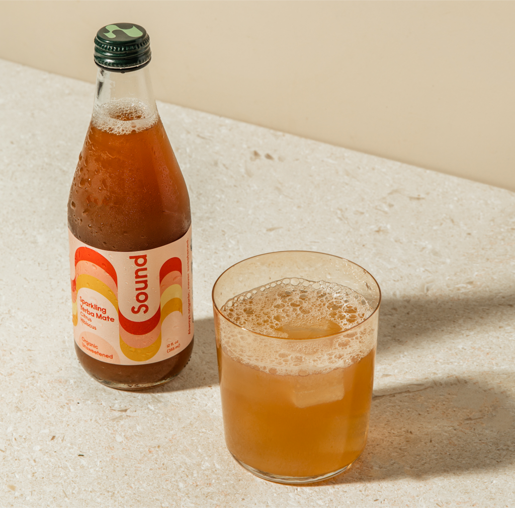 A bottle of Sound Sparkling Yerba Mate with a colorful wavy label sits on a light stone surface next to a glass filled with the bubbly golden beverage over ice.