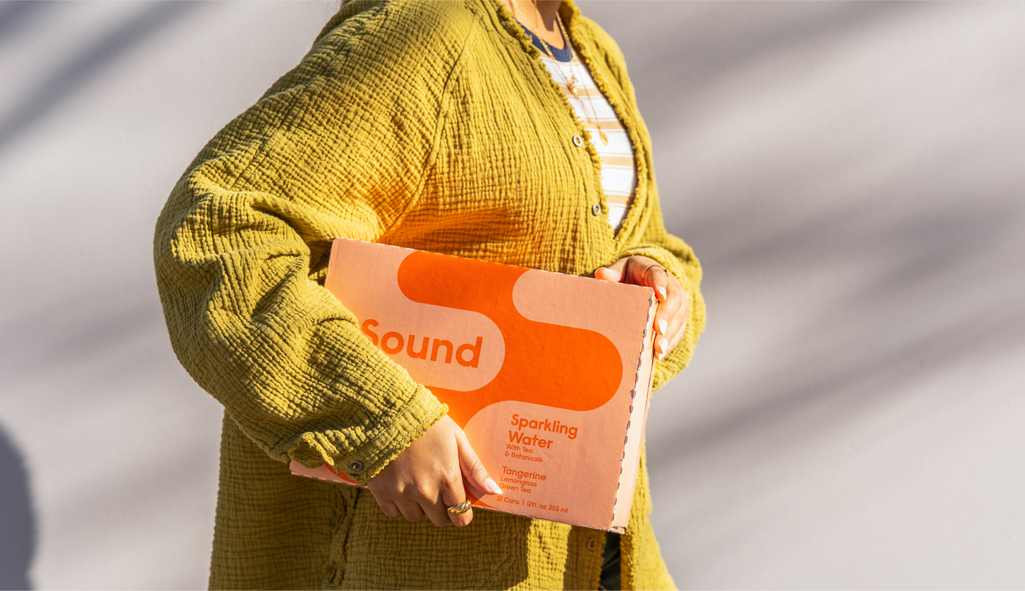 A person wearing a textured yellow cardigan holds a box of Sound sparkling water in tangerine flavor against a neutral background with soft shadows.