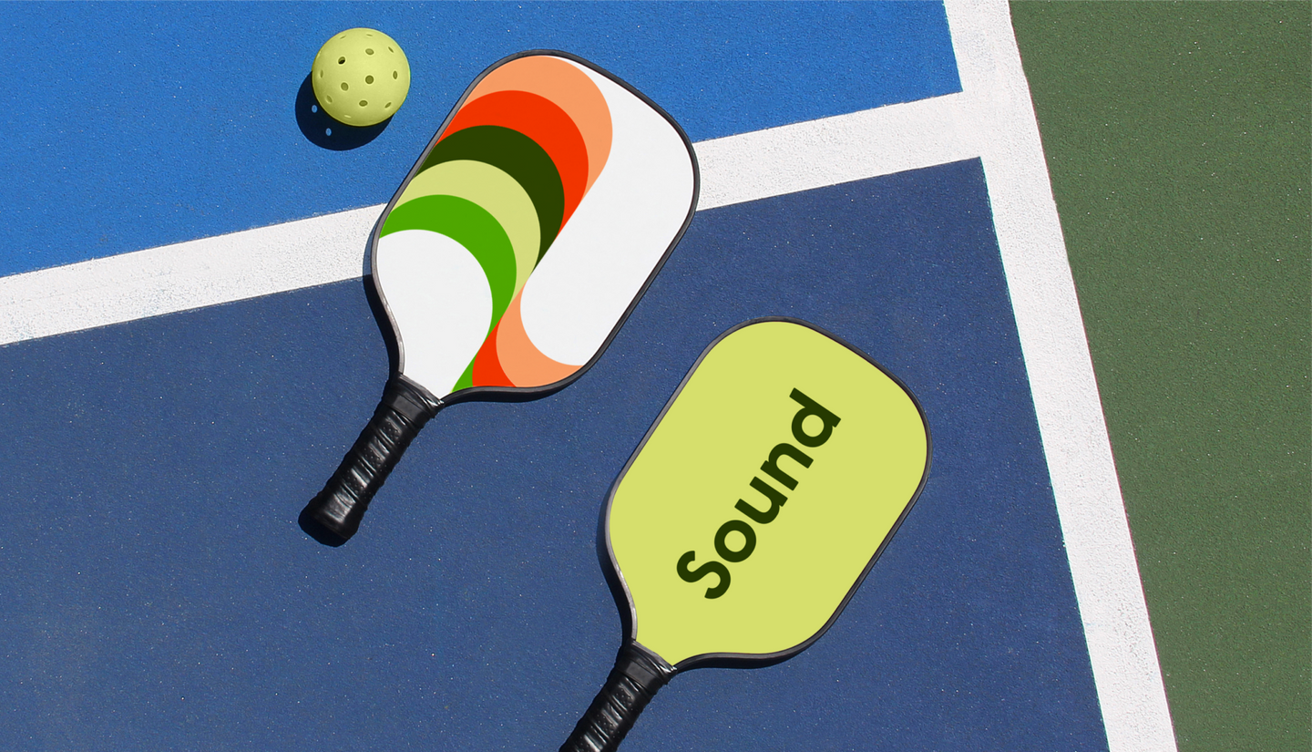 Two pickleball paddles with Sound branding and a yellow ball rest on a blue and green court near a white boundary line.