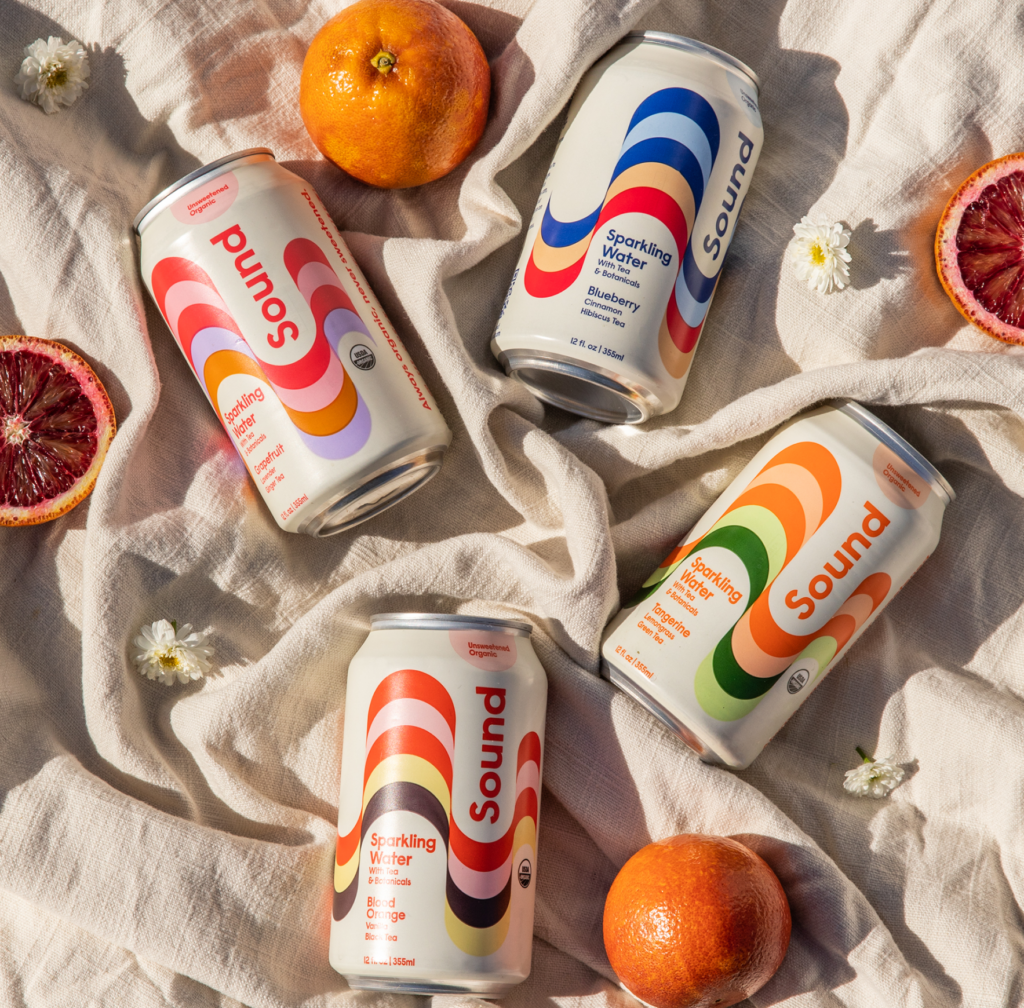 Four cans of Sound sparkling water are artistically arranged on a soft linen cloth, surrounded by fresh oranges, blood orange slices, and small white flowers.