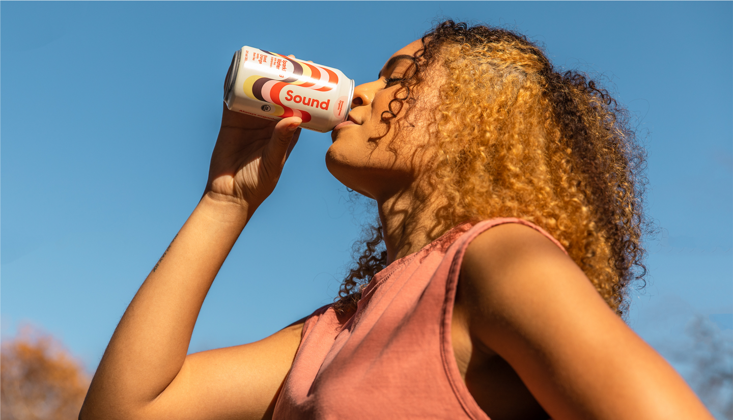 Young woman with curly hair drinks from a can of Sound sparkling water under a clear blue sky.