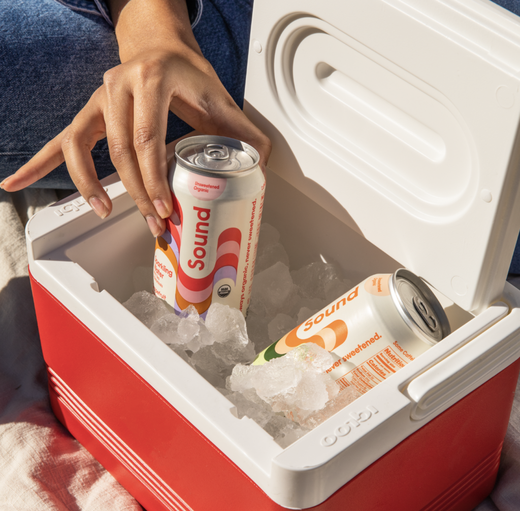 A person reaches into a red cooler filled with ice to grab a can of Sound sparkling water, with another can resting inside. The scene is set outdoors on a picnic blanket.