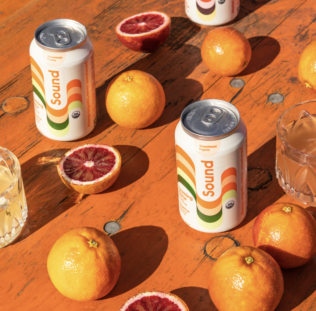 Cans of Sound sparkling water are arranged on an orange wooden table, surrounded by whole and halved blood oranges, along with glasses of citrus-infused drinks.