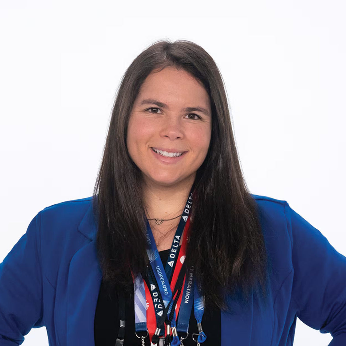 A smiling woman with long dark hair stands against a white background, wearing a bright blue blazer over a black top and multiple lanyards with event badges.