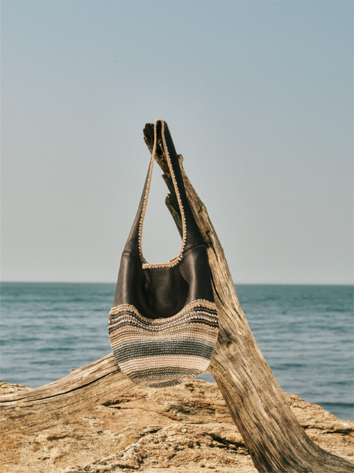 Boho-style woven and leather handbag hangs on a weathered driftwood branch against a serene ocean backdrop.