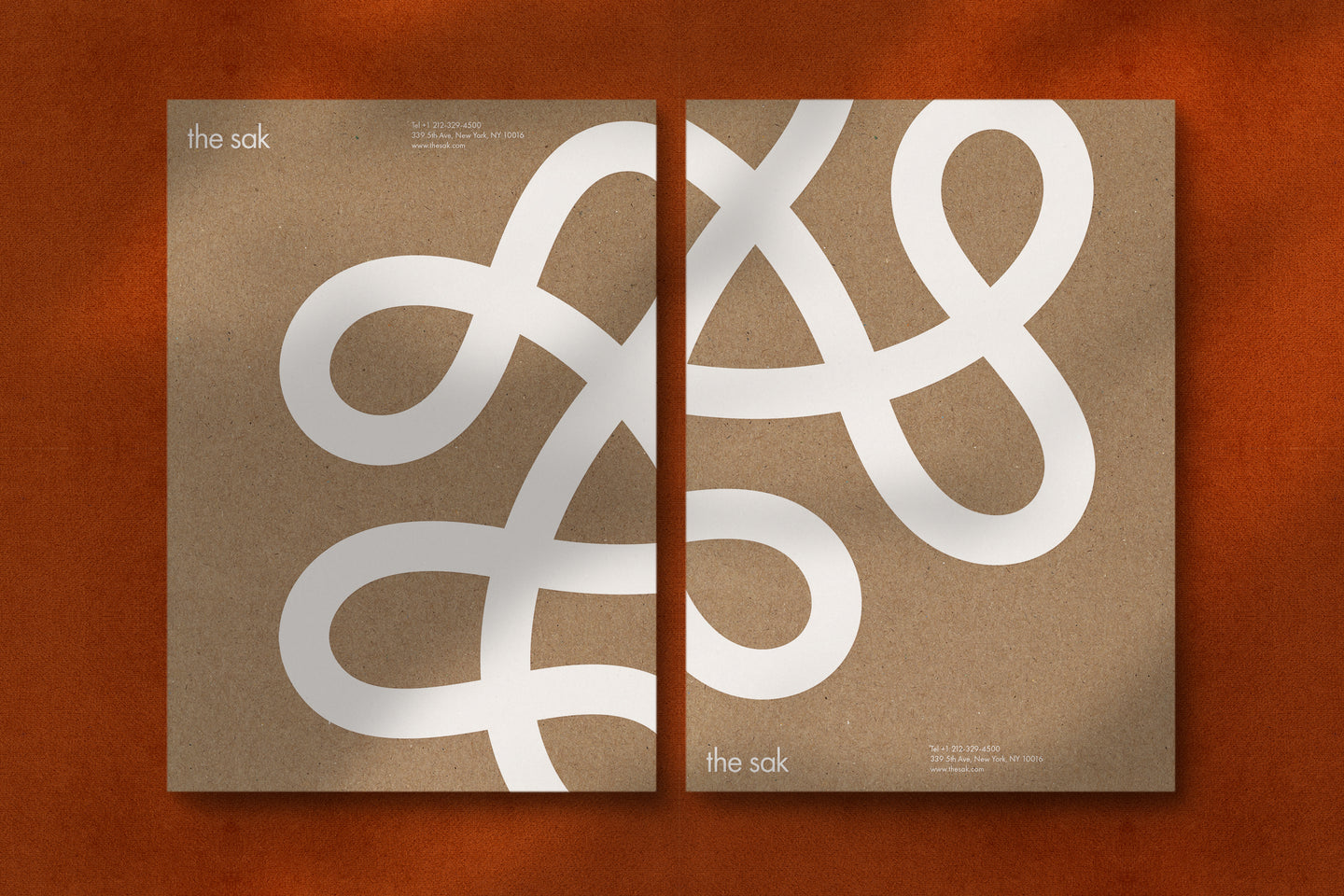 Two brown kraft paper folders featuring The Sak's signature white looping logo, placed side by side on a warm orange fabric background, creating a cohesive and natural brand aesthetic.