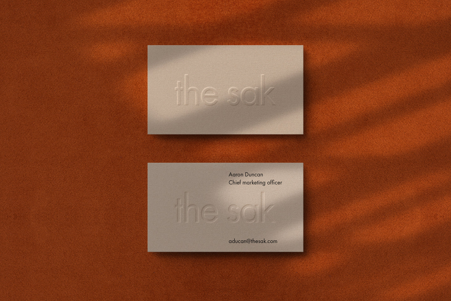 Two elegant business cards for 'The Sak' displayed on a warm, textured surface. One card features a subtle embossed logo, while the other includes contact details for Aaron Duncan, Chief Marketing Officer.