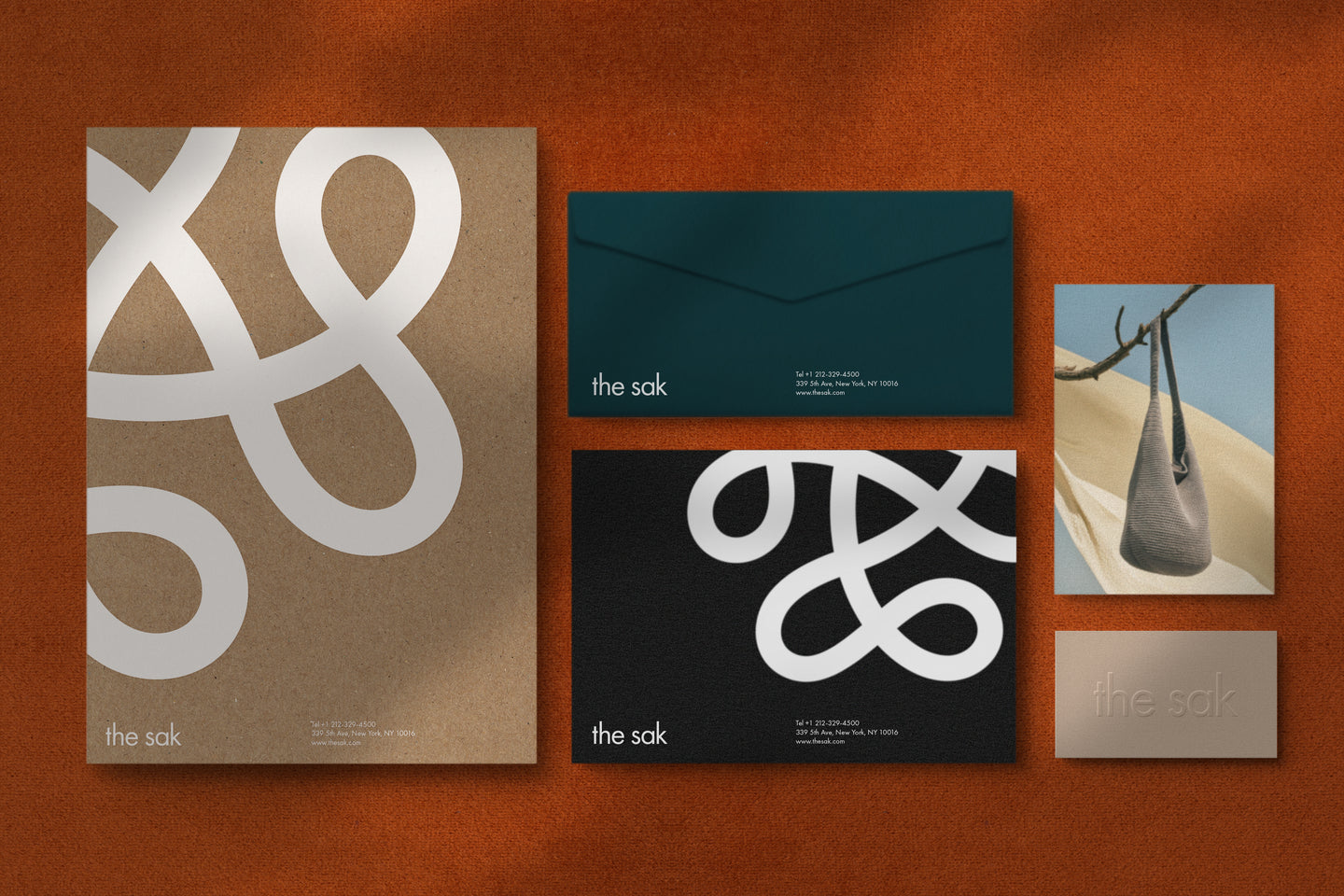 A collection of branded materials for The Sak displayed on a textured burnt-orange surface. Items include a kraft paper folder with a bold white abstract design, a dark green envelope, a black card with a similar white abstract pattern, a beige embossed business card, and an image of a woven handbag hanging from a tree branch. The materials emphasize a natural, sophisticated aesthetic.