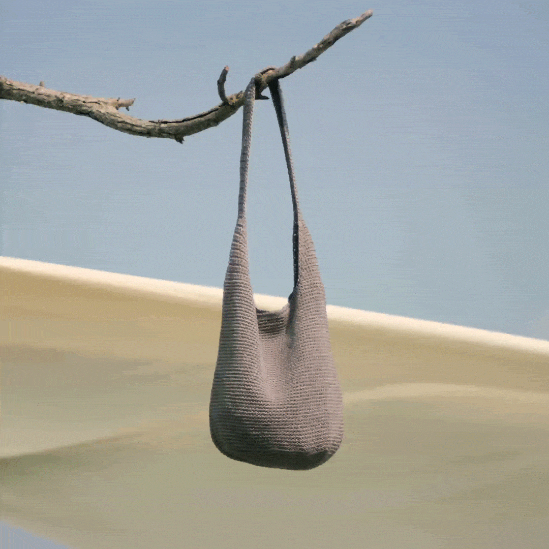 Knitted shoulder bag sways gently from a tree branch against a clear blue sky, with soft fabric billowing in the background.