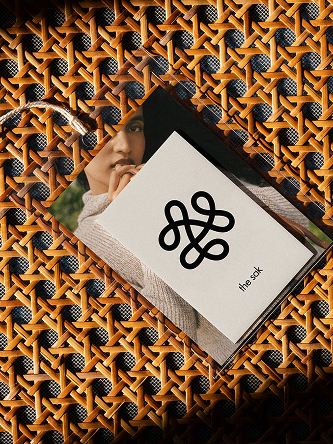 A branded card for The Sak featuring a bold black abstract logo and text, placed on a woven rattan surface. Beneath the card, a partially visible photograph of a woman in a beige sweater adds depth and texture to the composition. The design emphasizes natural materials and an artisanal aesthetic.