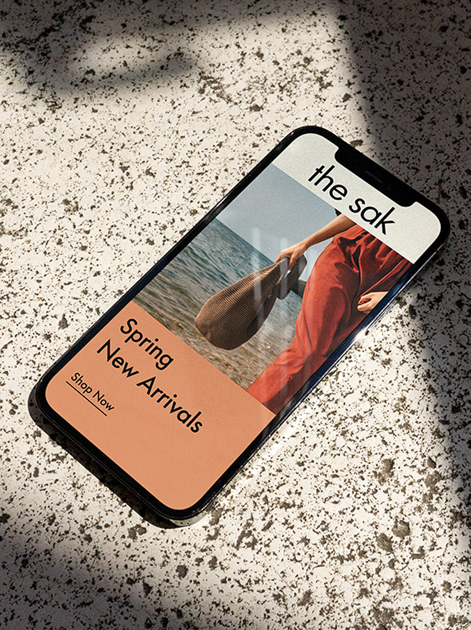 A smartphone displaying The Sak's Spring New Arrivals campaign, featuring a woman in a red outfit holding a woven bag by the beach. The ad includes a "Shop Now" button, set against a warm, earthy-toned design, resting on a textured stone surface with soft shadows.