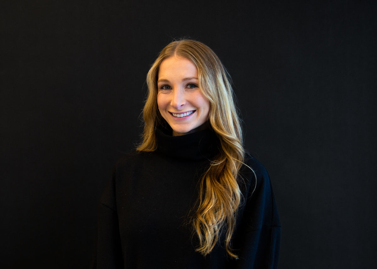 Portrait of a smiling woman with long, blonde wavy hair, wearing a black turtleneck sweater against a black background.