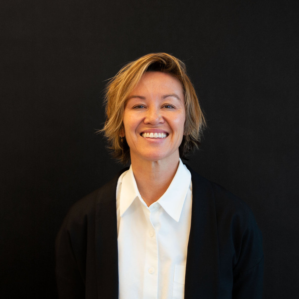 Portrait of a smiling woman with short, blonde hair, wearing a white collared shirt and black blazer against a black background.