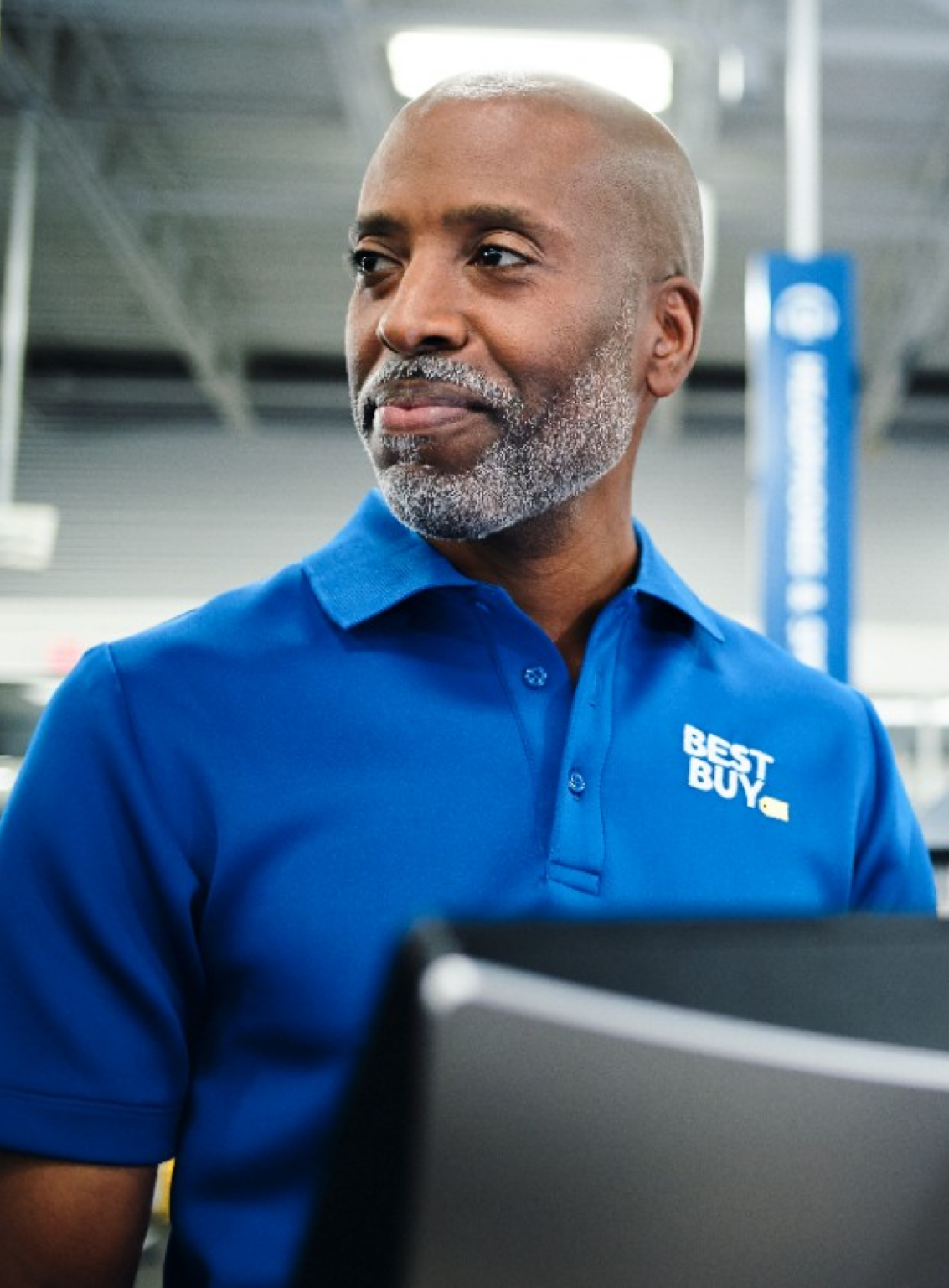 A Best Buy employee in a blue uniform stands in the store, looking off to the side with a confident expression.