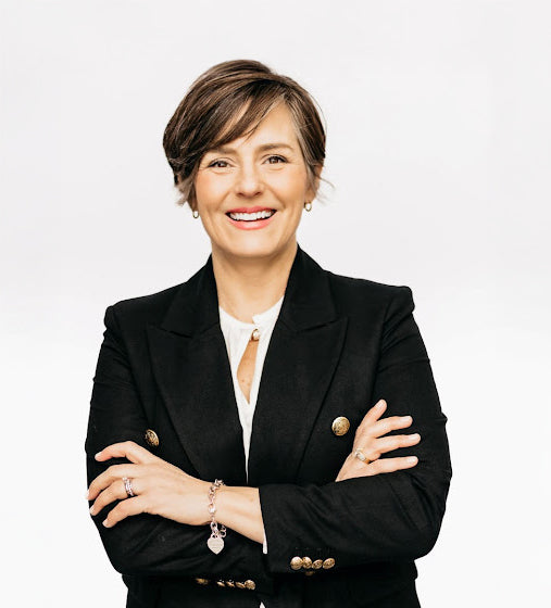 A confident person with short brown hair wearing a black double-breasted blazer with gold buttons, smiling with her arms crossed against a plain white background.