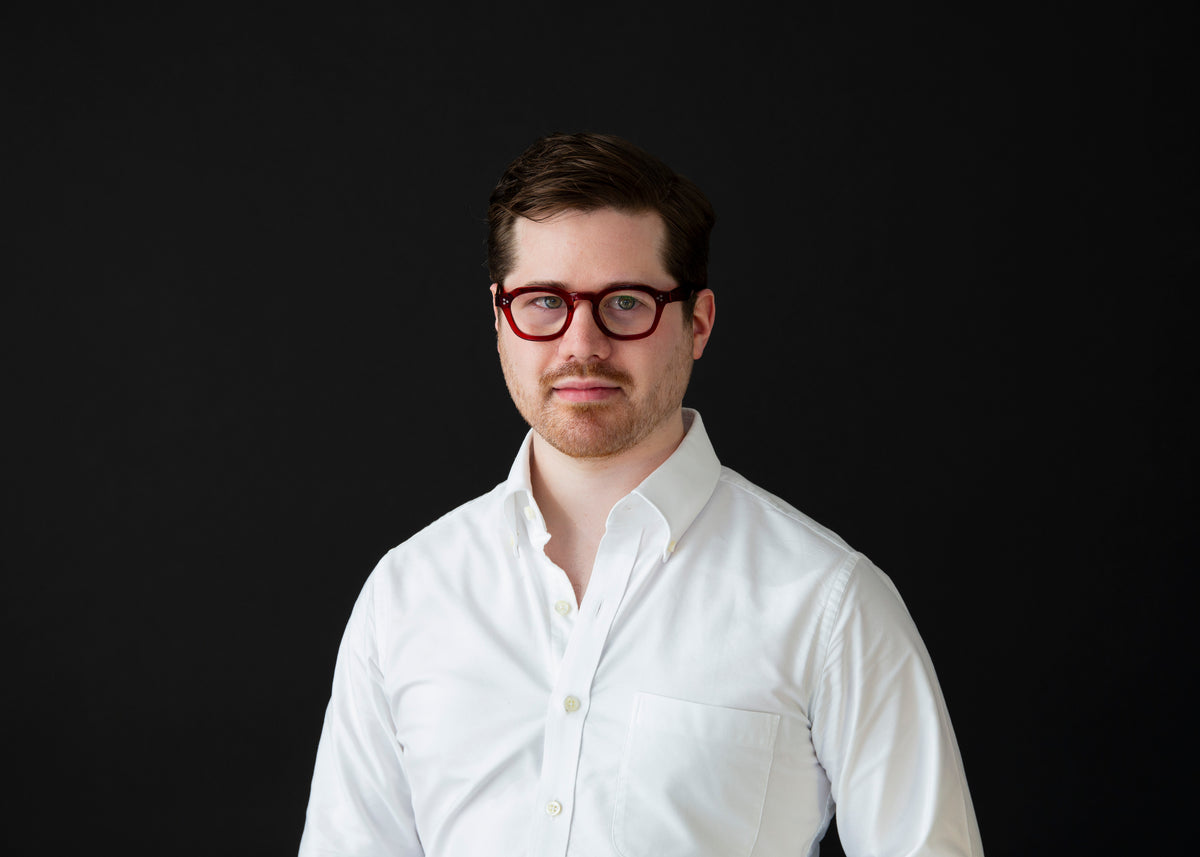 Portrait of a man with short brown hair, wearing red glasses and a white button-up shirt, against a black background.