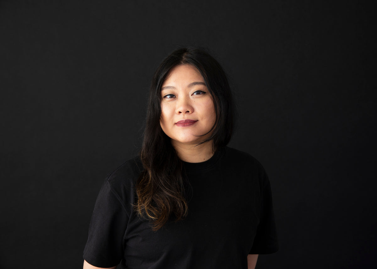 A portrait of a woman with long dark hair wearing a black t-shirt, posing against a solid black background with a confident, relaxed expression.