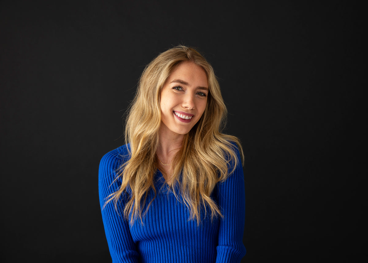 Portrait of a smiling woman with long, blonde wavy hair, wearing a bright blue sweater against a black background.